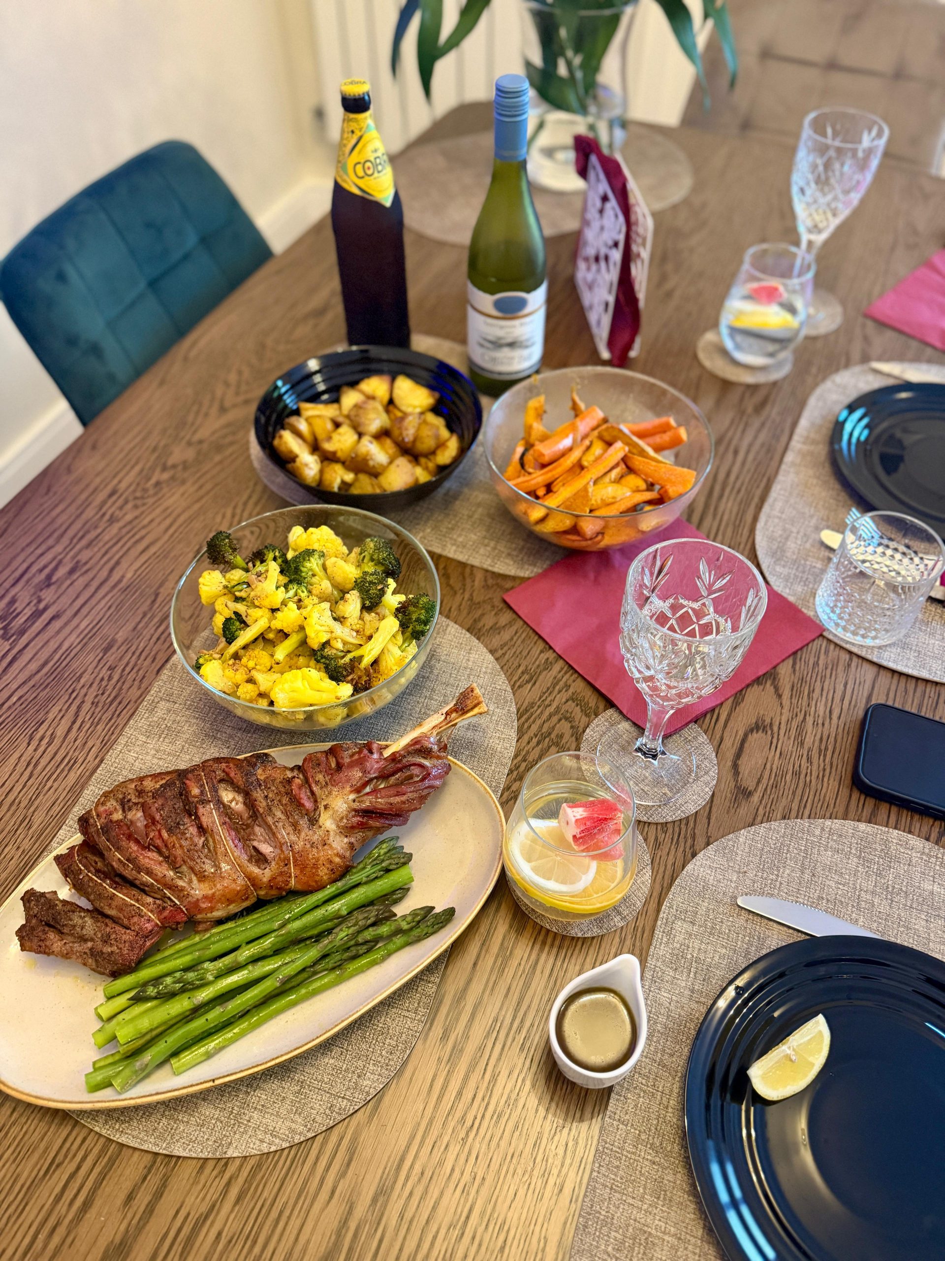 Full roasted mutton leg on a carving board surrounded by separate bowls of roasted asparagus, broccoli, sweet potato, cauliflower, and carrot.
