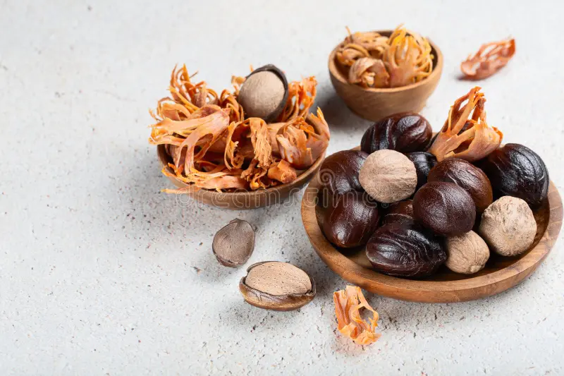 A wooden plate holds a cluster of smooth, dark-brown nutmeg seeds and a few beige mace blades; beside it, another bowl contains vibrant orange-red mace arils, all arranged on a light, textured tabletop.