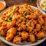 Crispy Bang Bang Chicken Bites coated in a creamy sweet and spicy sauce, garnished with fresh spring onions and served with coleslaw and dipping sauce on a rustic wooden table.