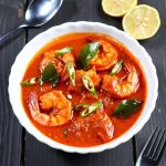 A vibrant bowl of Malabar prawn curry featuring large succulent shrimp in a rich orange-red coconut-based gravy, garnished with fresh cilantro and lemon slices, served in a white bowl on a rustic table with spoon, knife, fork, and warm candlelight in the background.