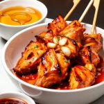 easy hot honey chicken tikka served in a white ceramic bowl, featuring glossy orange-red glazed chicken pieces on bamboo skewers, sprinkled with white sesame seeds and chopped green coriander, surrounded by a pool of shiny hot honey sauce
