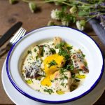 Marry Me Chicken: tender seared chicken pieces in a vibrant creamy sauce with sun-dried tomatoes, topped with chopped fresh parsley and a drizzle of olive oil. The dish is surrounded by small bunches of wild rosemary, thyme, lavender flowers, and other fresh herbs arranged on a rustic wooden table next to a silver fork and knife.