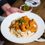A vibrant, mouth-watering 3D photographic close-up of creamy butter chicken pasta served by a professional chef. The rich, orange-colored sauce-coated pasta is garnished beautifully and presented on a stylish plate on a dining table, accompanied by side dishes like naan bread and salad, with a fork and knife elegantly placed beside it. Bright, appetising colours and professional food styling.