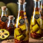 Glass bottles filled with traditional Monkey Jackfruit Pickle, showing tender jackfruit cubes immersed in golden mustard oil with red chilli flakes and spices. The bottles are displayed on a rustic wooden surface with a sliced monkey jackfruit in the background