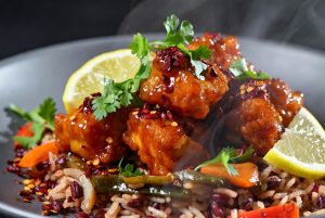 image of crispy glazed bite-sized pieces served over a colorful red and black rice blend with sautéed vegetables, garnished with fresh coriander, red chili flakes, and lemon wedges, with visible steam rising from the hot dish.