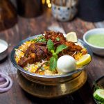 Hot Bhatkali biryani served in a brass vessel with chicken, boiled egg, fried onions, mint, lemon, accompanied by raita, chutney, papad, and salad on a rustic dining table.