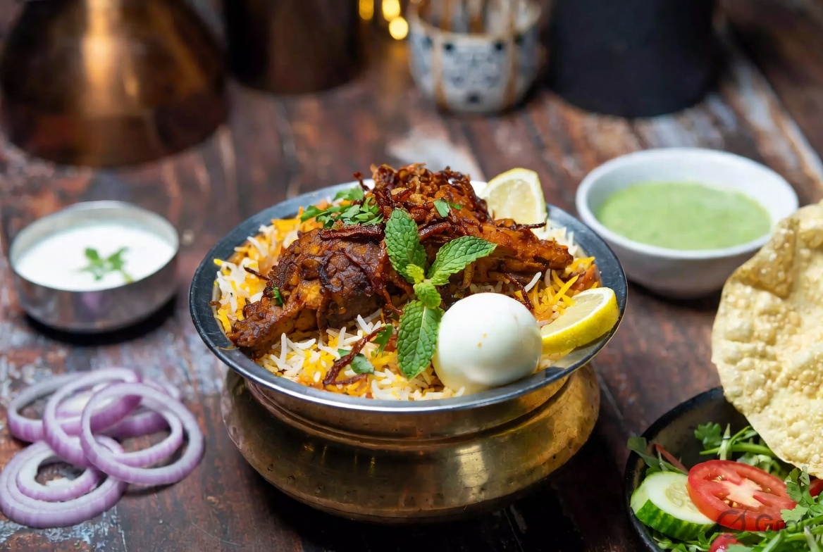 Hot Bhatkali biryani served in a brass vessel with chicken, boiled egg, fried onions, mint, lemon, accompanied by raita, chutney, papad, and salad on a rustic dining table.