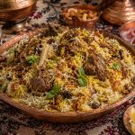 Authentic Kashmiri Mutton Biryani served on a traditional dastarkhan in a copper platter with long-grain saffron basmati rice, tender bone-in mutton, fried onions, mint, raisins, cashews, yoghurt raita, onion salad, lemon wedges, and Kashmiri accompaniments, with the madhusavara.com watermark on the right.