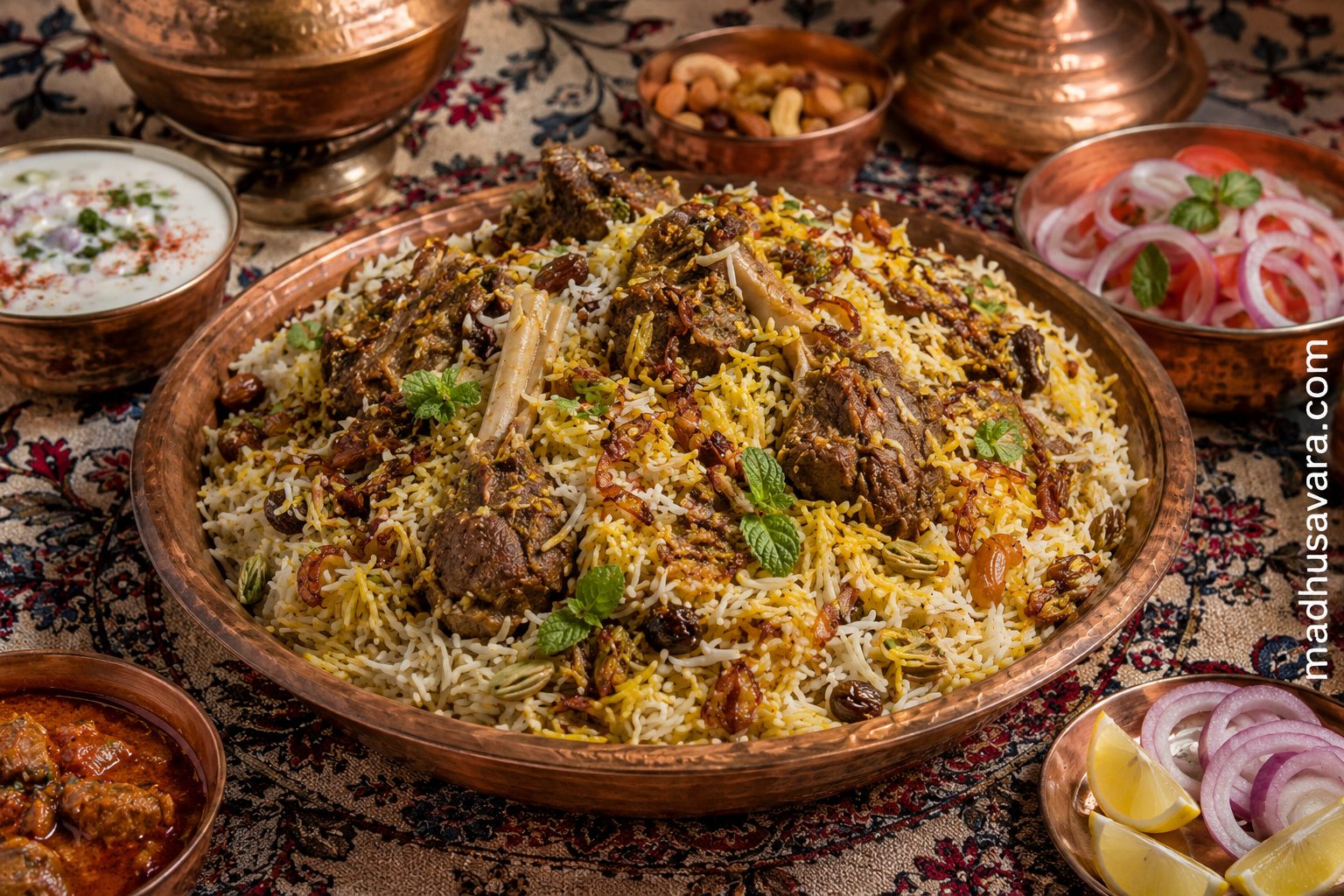 Authentic Kashmiri Mutton Biryani served on a traditional dastarkhan in a copper platter with long-grain saffron basmati rice, tender bone-in mutton, fried onions, mint, raisins, cashews, yoghurt raita, onion salad, lemon wedges, and Kashmiri accompaniments, with the madhusavara.com watermark on the right.