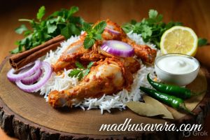 Premium basmati rice, fresh chicken pieces, sliced onions, mint leaves, coriander, spices, and yogurt on rustic wood with madhusavara.com watermark.