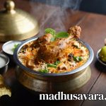Authentic Malabar Chicken Biryani served in traditional brass handi on a wooden table, garnished with fried onions, cashews, mint and accompanied by raita, boiled egg, lemon and papad - vibrant Kerala style Thalassery biryani