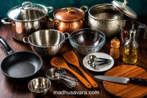 Traditional cooking utensils for preparing Sindhi Biryani including a copper biryani pot, stockpot, colander, mixing bowls, measuring spoons, chopping board, knife, and wooden spatula.