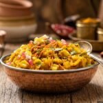 Corn bhel chaat served with a silver spoon in a traditional copper bowl on a wooden table, featuring sweet corn kernels, sev, pomegranate seeds, and fresh cilantro garnish.