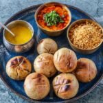 Traditional Madhya Pradesh Dal Bafla thali featuring boiled and baked baflas, a bowl of spicy tuar dal topped with fresh cilantro, sweet crumbled churma, and a hammered katori of pure liquid ghee served on a blue ceramic platter.